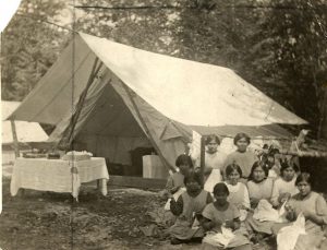 一群青年员工坐在大帆布帐篷旁缝纫, 前面有张表,后台有树