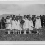 一组六位青年站在白毕业礼服上持有花束,大群穿制服站在身后排队